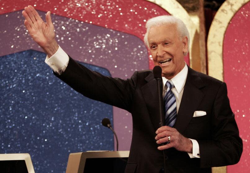Bob Barker in a black suit and striped tie holding a microphone in his right hand and holding out his left arm