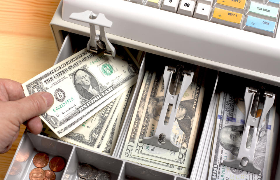 A person's hand is placing a one-dollar bill into a cash register drawer filled with various denominations of paper money and coins