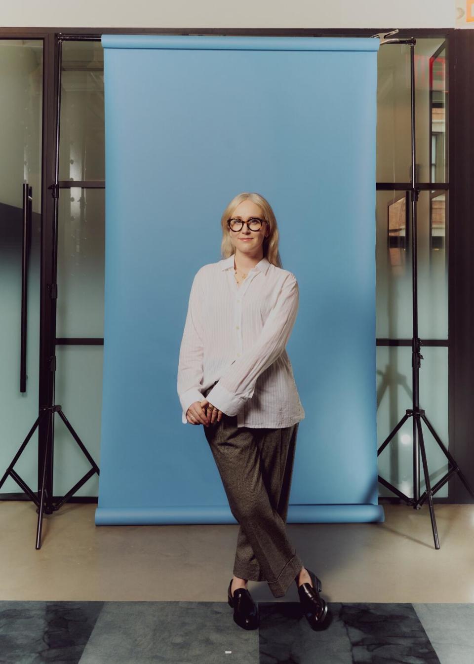 A blond woman wearing glasses, a white button-down shirt and gray trousers, poses in front of a sky blue backdrop.