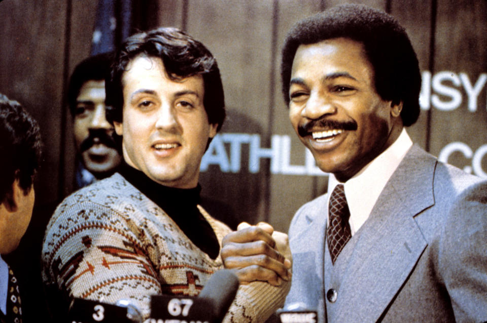 Sylvester Stallone and Carl Weathers smiling and shaking hands at a press event. Stallone is wearing a patterned sweater, and Weathers is in a gray suit