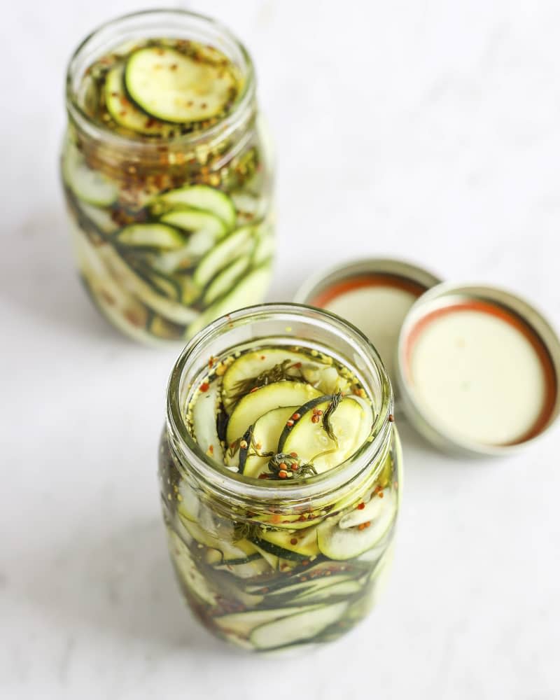 A photo of two mason jars with pickled zucchini slices in them, as seen from the top down, and the lids in the background.