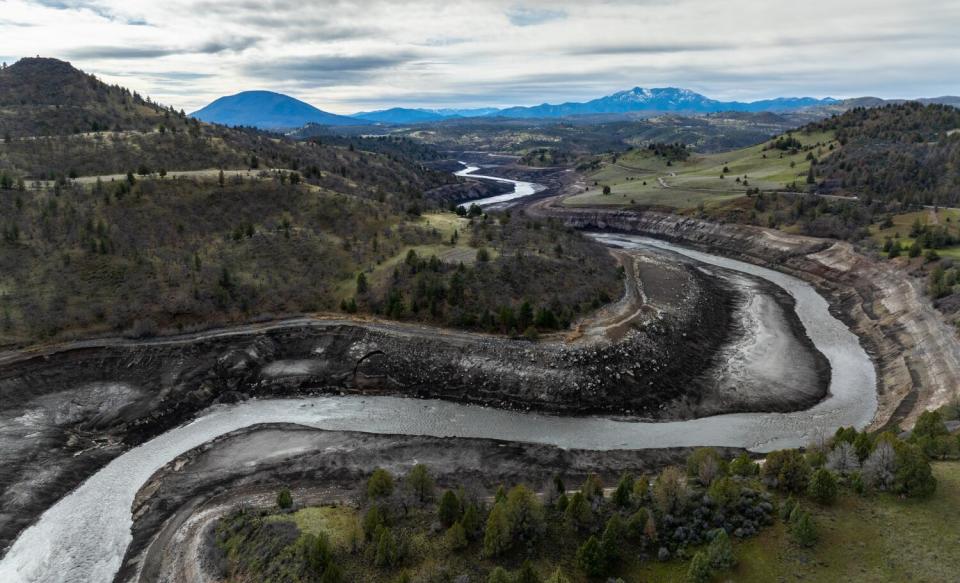 A river curves back and forth as it flows through layered sediment amid green hills and trees, with mountains in the distance