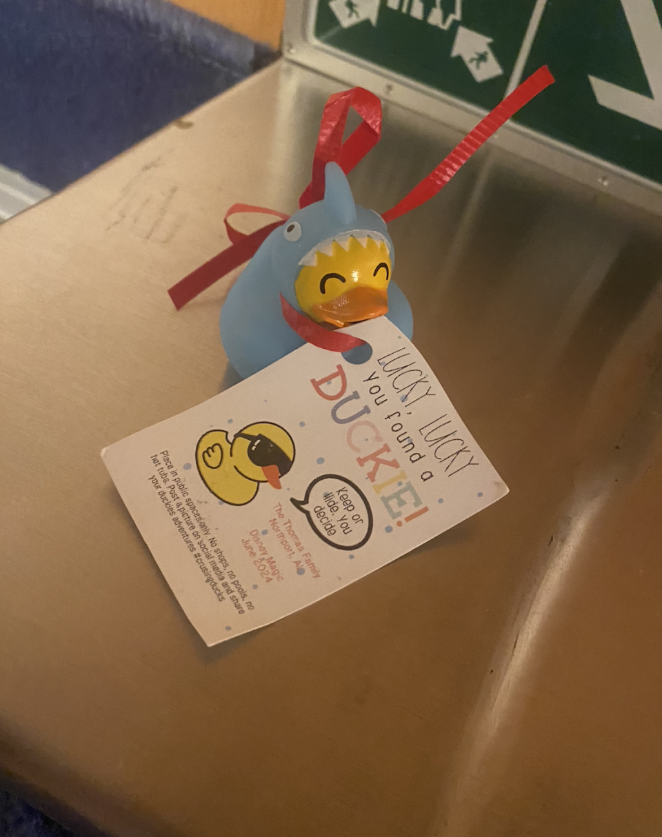 A small blue rubber duck wearing a shark costume with a red ribbon around its neck, holding a card that reads, "Lucky Lucky, you found a DUCKIE!"