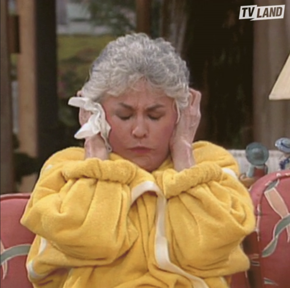 Character from "The Golden Girls" wearing a bathrobe, looking annoyed with hands on ears