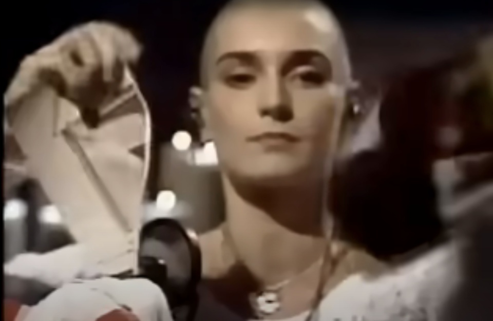 Sinead O'Connor, with a shaved head, tearing a photograph while wearing a white outfit on stage