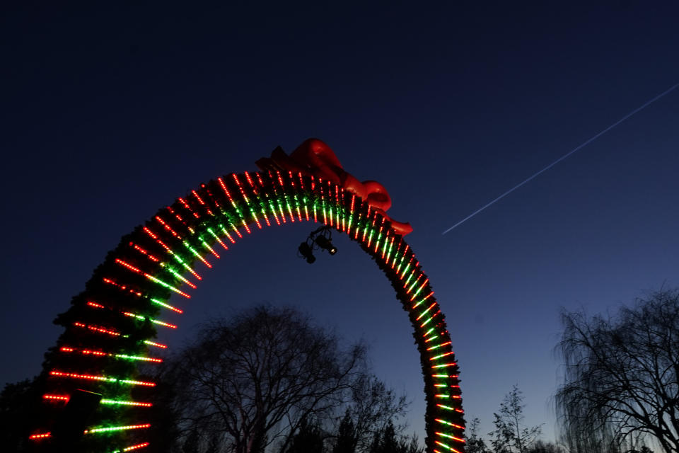 A commercial air plane streaks across the the post-sunset sky above the "Welcome Arch" at the beginning of a 1.3-mile path through the Chicago Botanic Garden's fifth annual Lightscape holiday experience of light and music in Glencoe, Ill., on Thursday, Dec. 14, 2023. (AP Photo/Charles Rex Arbogast)
