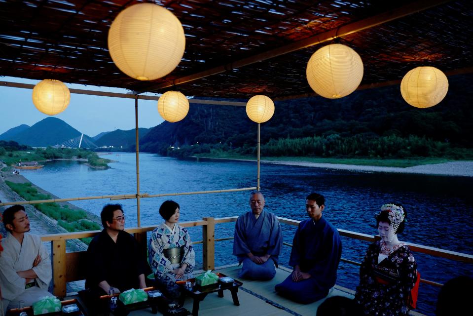 A group of people in robes sit on their knees on an observation deck on a river.