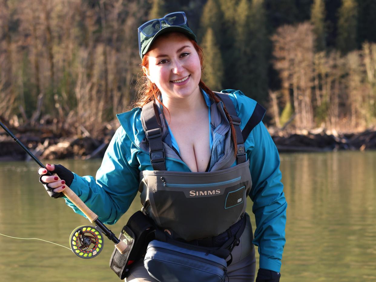 OnlyFans creator Isla Moon standing in a lake surrounded by forest tress. She wears grey and blue waterproof clothing and holds a fishing rod in her right hand.