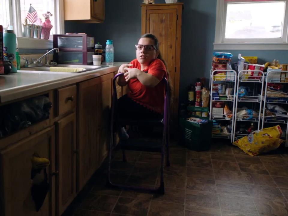 Natalia Grace stands on a step stool near the kitchen sink in a red t-shirt and glasses.