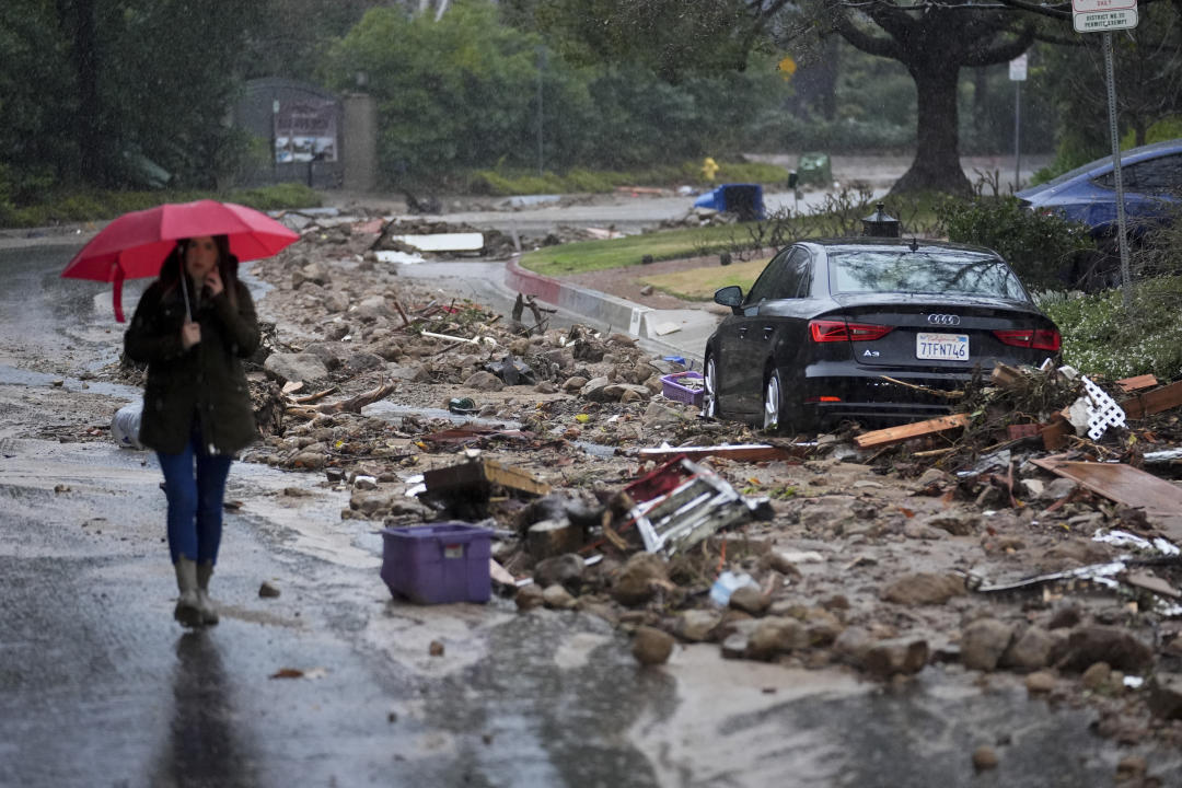 Mud and debris is strewn along the side of a road as a woman walks next to it holding a red umbrella.