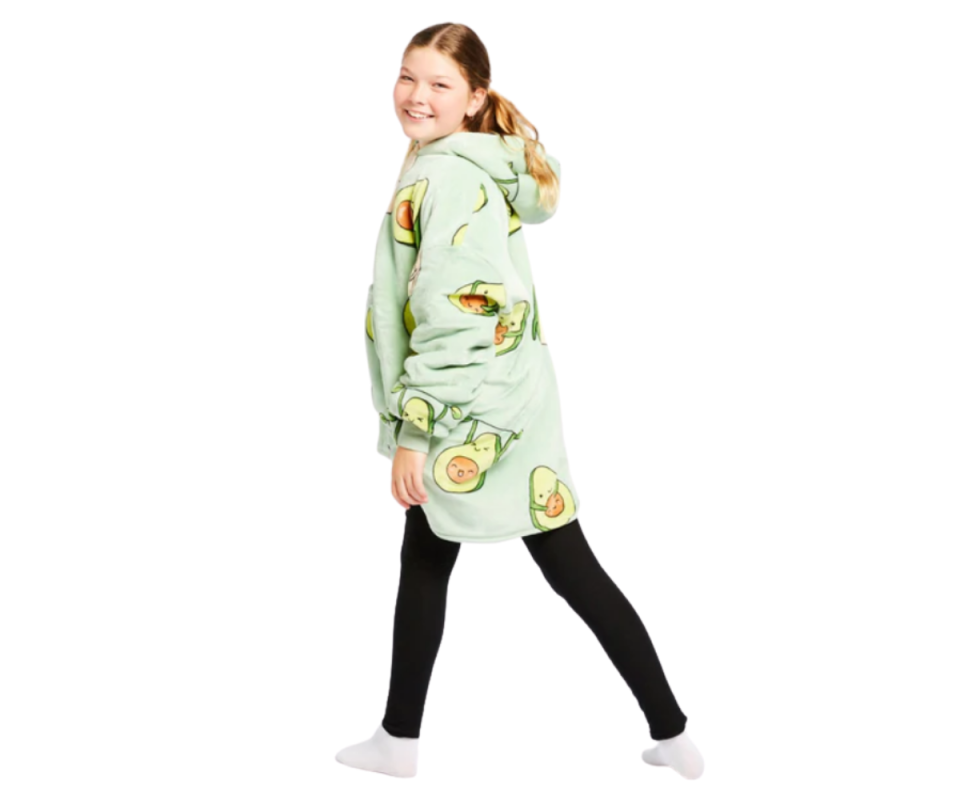 A caucasian blonde girl wears a pale green Oodie with cartoon Avocados on it with black leggings on a white background.