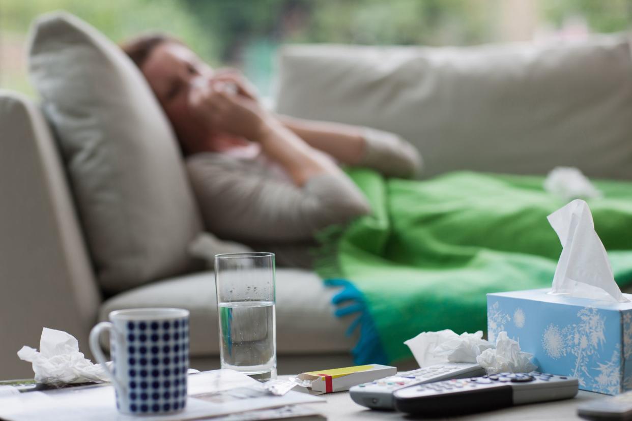 Foreground of table with tissue box, used tissues, medicine, and water. Background of a sick women laying on a couch under a blanket and blowing her nose.