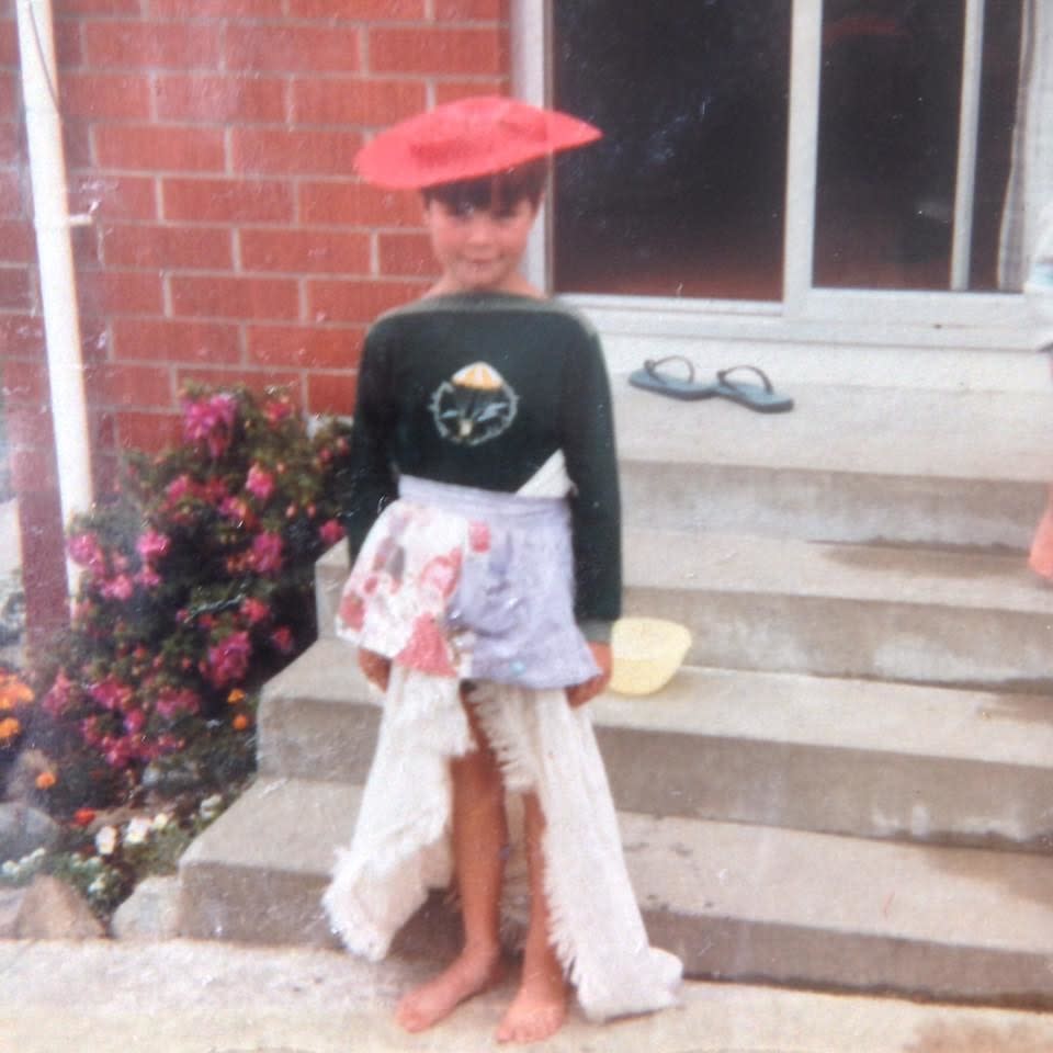 A barefoot child standing outside a house waring an apron dress and a disc hat