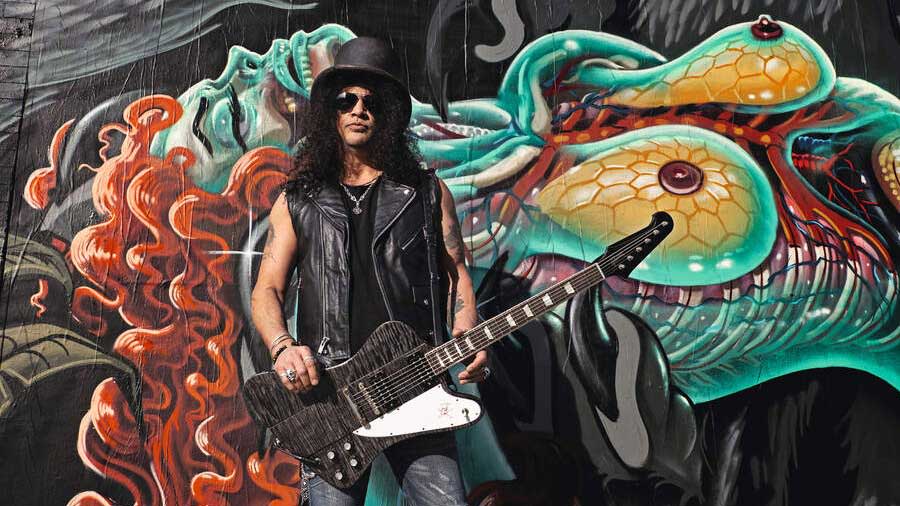  Slash standing against a grafitti'd wall with a guitar. 