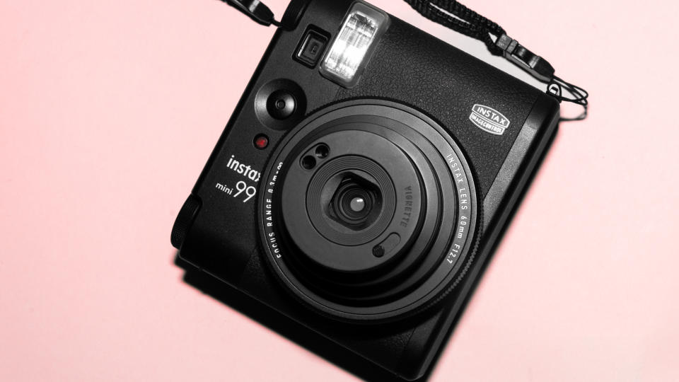 A photograph of the Fujifilm Instax mini 99 in black, set against a pink background.