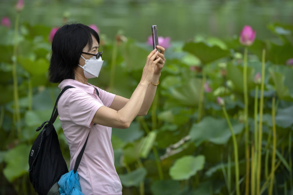 A woman wearing a face mask takes photos of lotus flowers at a public park in Beijing, Tuesday, June 28, 2022. (AP Photo/Mark Schiefelbein)