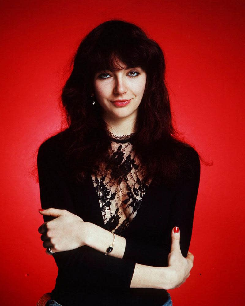 A formal portrait of Kate Bush at the height of her fame, arms folded and dressed in black against a red background