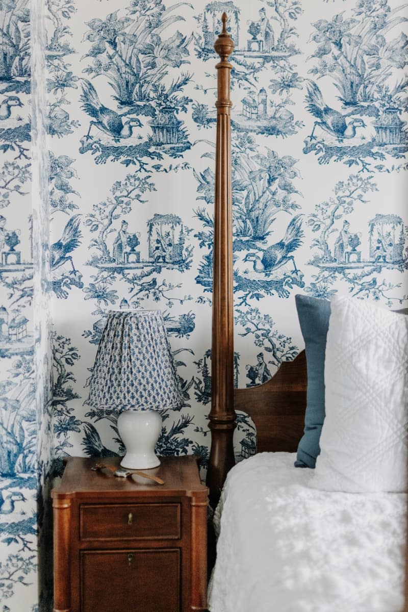 Blue toile wallpaper, small dark wood nightstand with pleated blue lamp shade, wood four poster bed