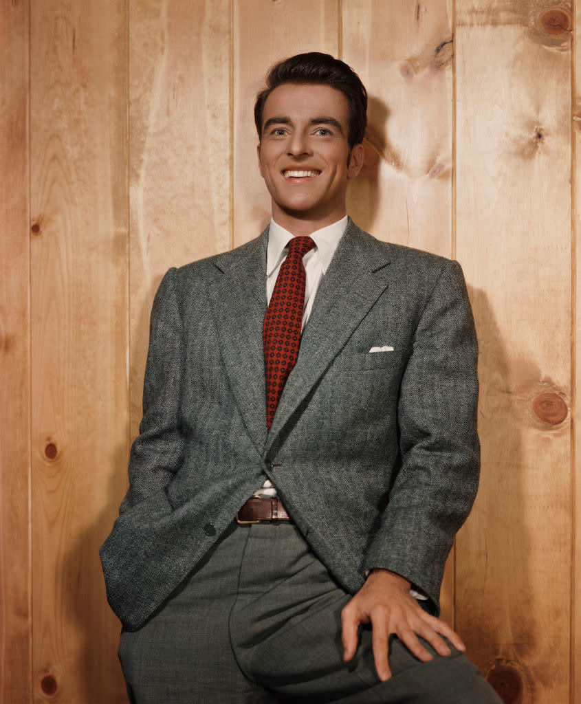 A man in a suit and tie leans casually against a wooden wall, smiling confidently