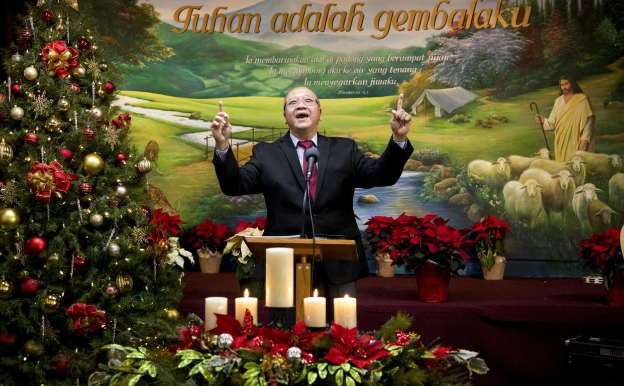 A man at a podium smiles and points to the sky, next to a Christmas tree