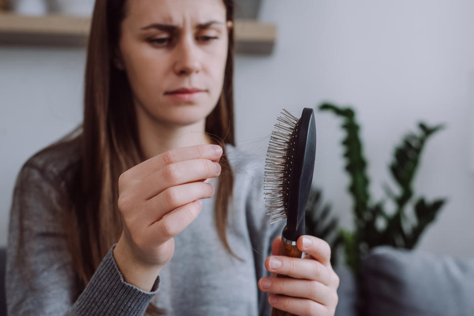 Selective focus of young girl sitting on sofa brushing hair with hair tool, worrying about hair loss shedding or bad condition. Upset brunette woman styling hair with comb, hormonal imbalance concept