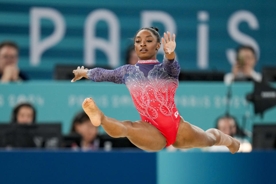 Simone Biles performs a mid-air leap during a gymnastics competition in Paris. She is wearing a sparkling athletic outfit