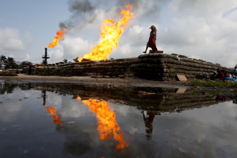 A reflection of two gas flaring furnaces and a woman walking on sand barriers is seen in the pool of oil-smeared water at a flow station in Ughelli, Delta State