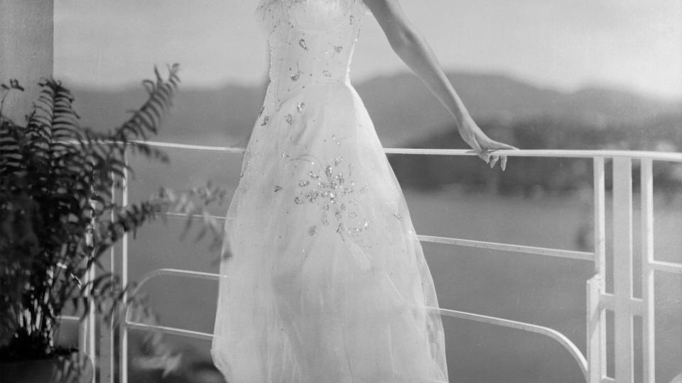 rita hayworth smiiles and stands with her back to a balcony railing that she holds with both hands, she wears a white strapless dress