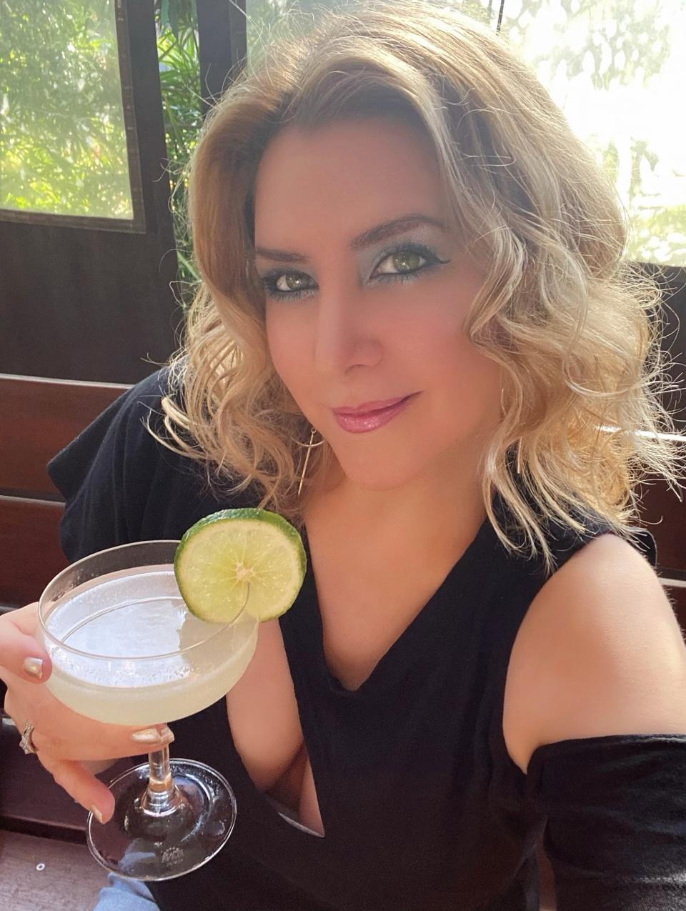 Woman with wavy blonde hair holding a cocktail with a lime garnish, smiling at the camera