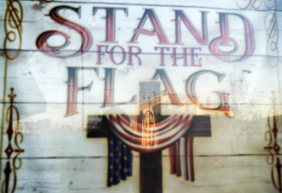 A mural of the Virgin Mary, painted on the side of a building, is reflected in a poster which reads "Stand for the Flag, Kneel for the Cross," outside a church in Roseville, Mich., Saturday, Oct. 31, 2020. As the traditional Election Day closes in, Americans are exhausted from constant crises, on edge because of volatile political divisions and anxious about what will happen next. (AP Photo/David Goldman)