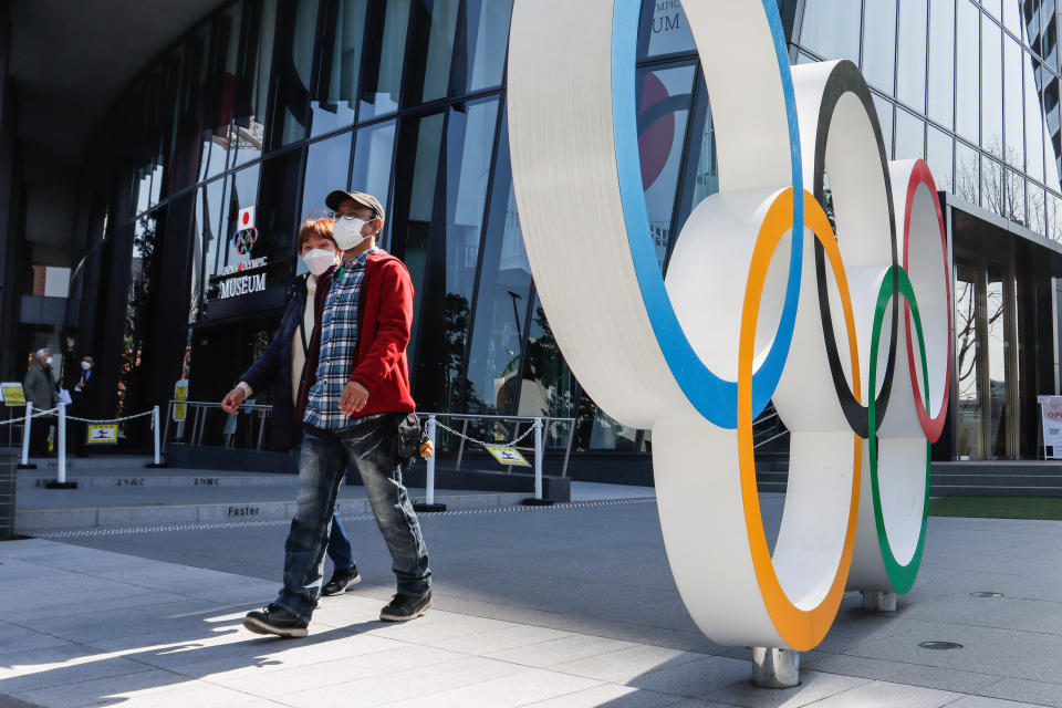 TOKYO, JAPAN - 2021/03/11: People wearing facemasks are seen behind the Olympic symbols of the five interlaced rings near the National Stadium in Tokyo.
About 80% of Japanese in recent polls say the Olympics should be postponed or cancelled, and almost as many do not want fans from abroad. (Photo by James Matsumoto/SOPA Images/LightRocket via Getty Images)