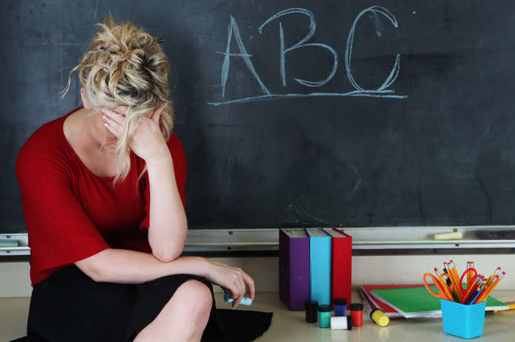 Elementary classroom setting with tired or frustrated teacher holding her head. She's sitting in front of an chalkboard with ABC