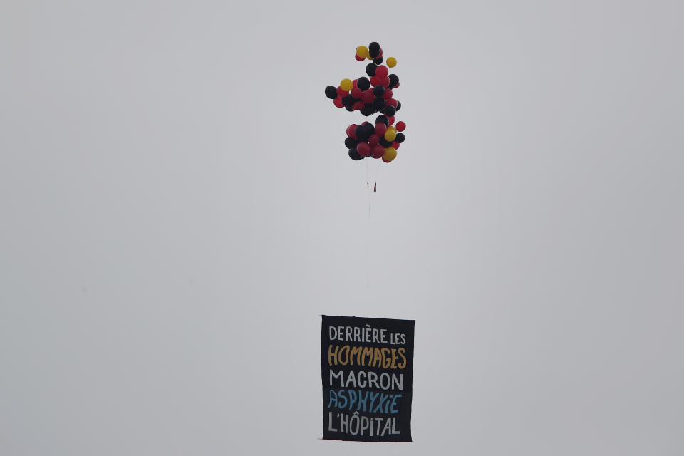 A placard suspended from ballonns reads « Behind the tributes, Macron asphyxiates the hospital » during the Bastille Day military parade, Tuesday, July 14, 2020 in Paris. France are honoring nurses, ambulance drivers, supermarket cashiers and others on its biggest national holiday Tuesday. Bastille Day's usual grandiose military parade in Paris is being redesigned this year to celebrate heroes of the coronavirus pandemic. (AP Photo/Francois Mori)