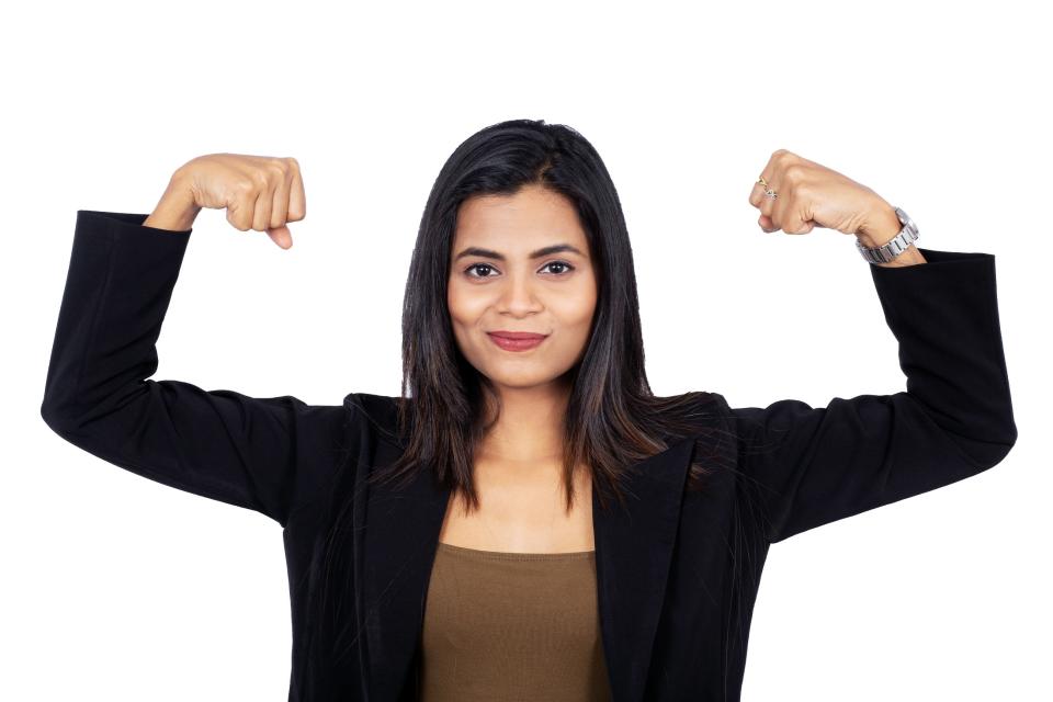 Portrait of confident Indian businesswoman flexing her muscles to show her girl power. Beautiful female professional is over white background. She is wearing elegant suit.