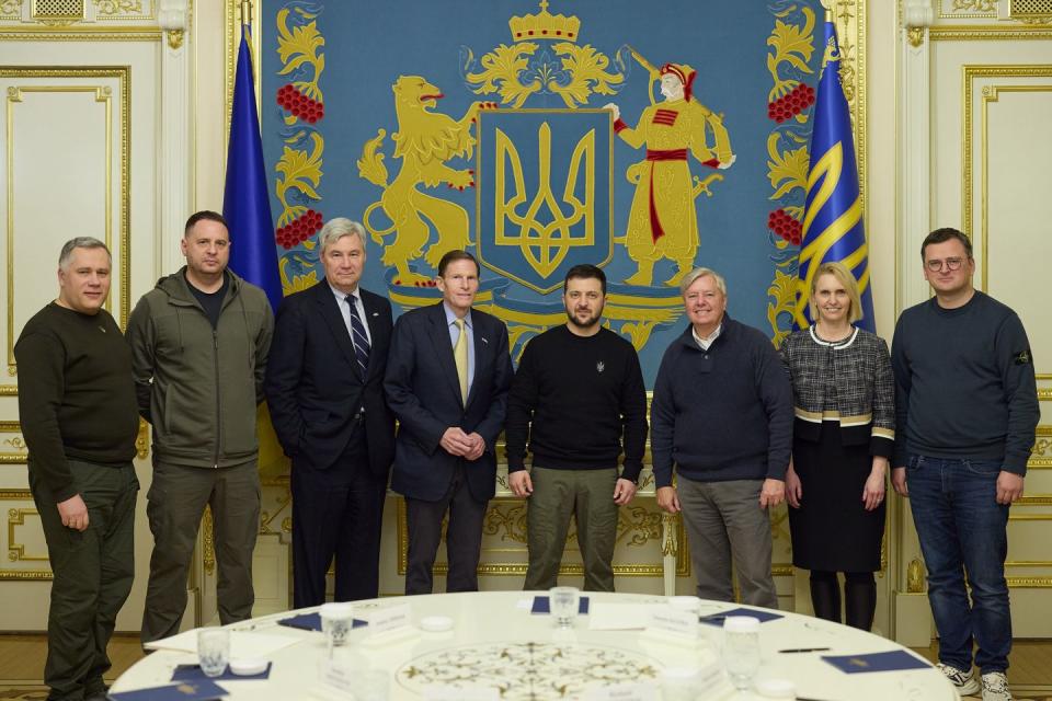 a group of people stande side by side behind a table, in front of a ukranian banner and flags