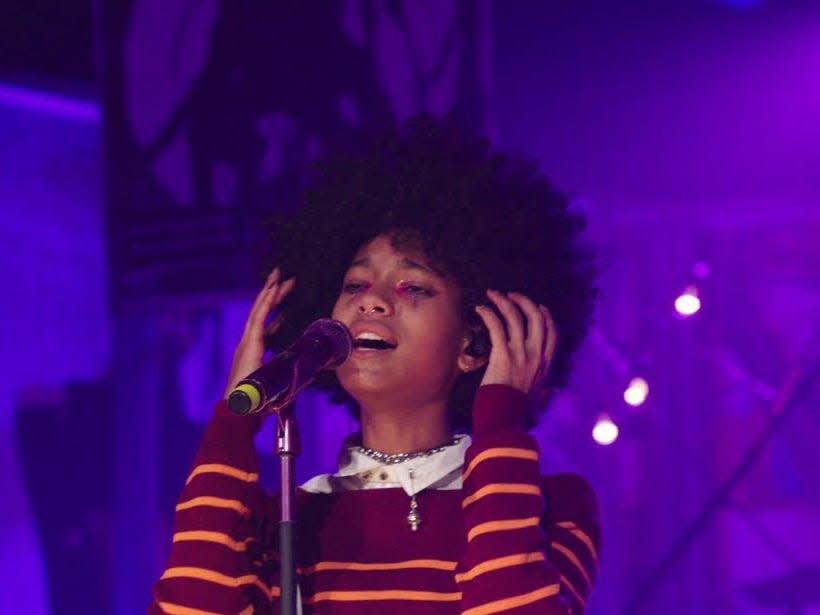 WILLOW is in a white collared shirt layered with an orange and maroon striped sweater. She has both hands near her ears, as she sings in the mic. She has an afro.