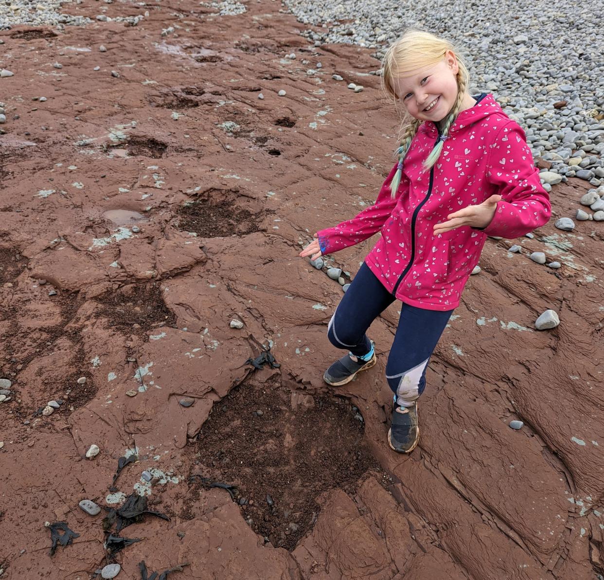 A blond girl in brains motions toward dino footprints on the beach