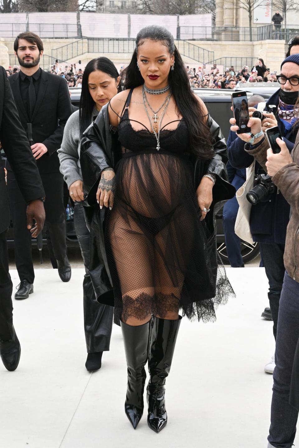 Rihanna in a black sheer mesh dress revealing a bra and underwear underneath. She has a black leather jacket worn off-the-shoulder and has knee-high leather pointed boots.
