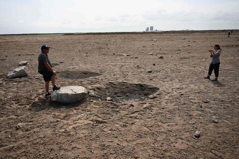 woman takes photo of man standing with a foot on a large chunk of concrete next to a crater in a dry dirt field