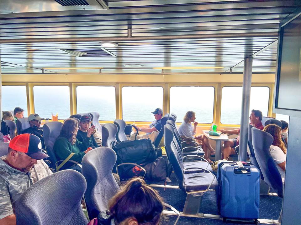 Several black cushioned seats, many of them filled with passengers or luggage, on a ferry. Windows looking out at the ocean sit behind the passengers.