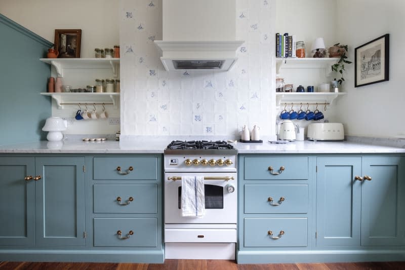 Blue cabinets in newly renovated kitchen with blue and white tiles on backsplash with small vintage inspired gas stove. Opened shelves on either side of exhaust.