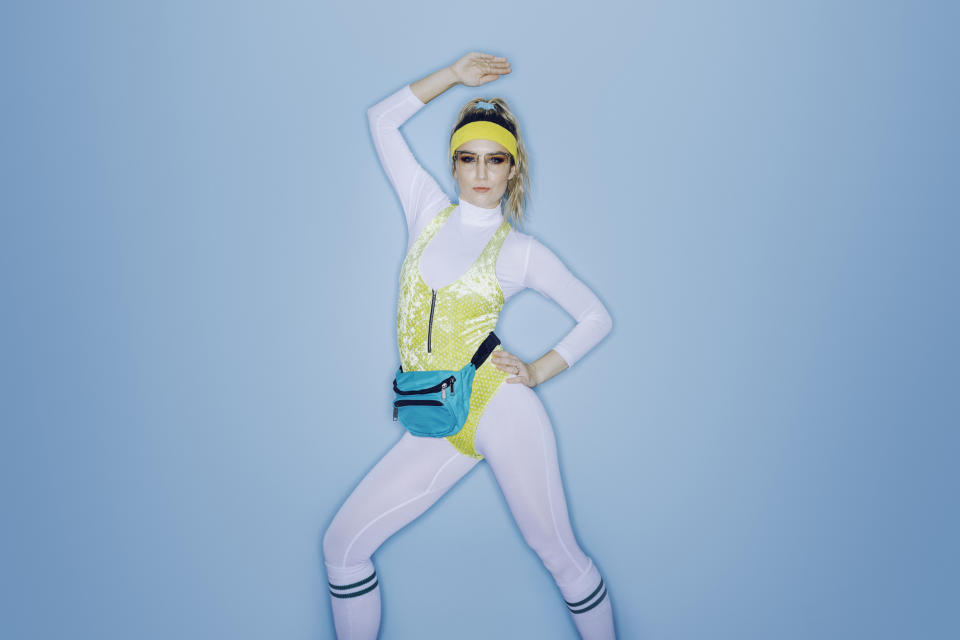 Woman in a retro workout outfit posing confidently. She's wearing a yellow headband, blue fanny pack, white leotard, and yellow lace unitard. Her arm is raised