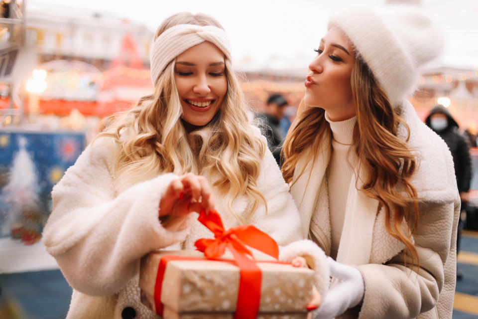 Two smiling female girlfriends in hats and warm coats are walking and laughing on a decorated street in Europe on Christmas day. cold weather outdoor. A loving couple of two lesbians celebrate the new year together congratulate each other and give gift boxes