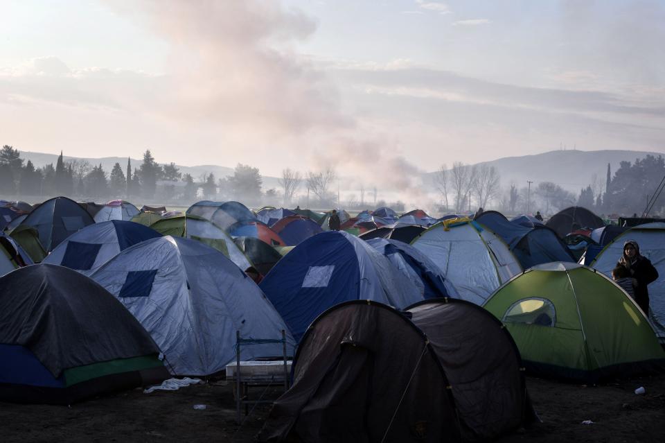 Smoke rises in the distance behind a woman standing among tents in the makeshift camp in Idomeni. Since the border crossings have slowed, up to 12,000 people are stuck in the camp.&nbsp;