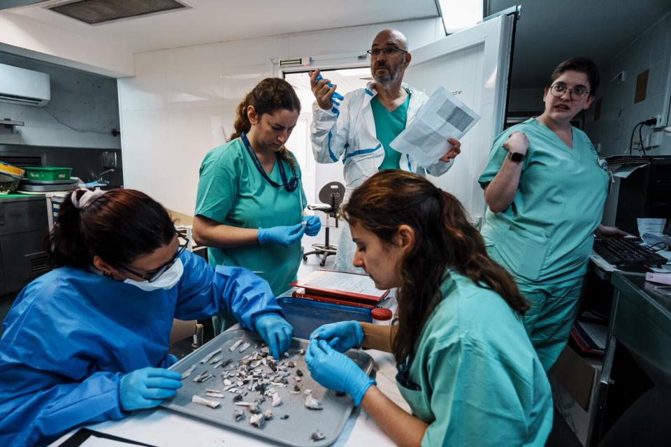 Two women in blue scrubs and gloves sort through fragments on a tray as three other people in scrubs stand nearby