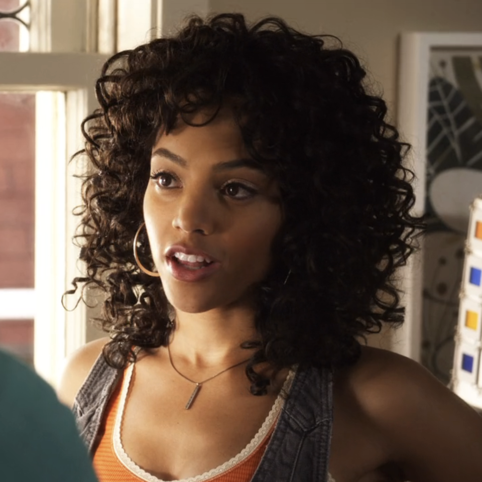 Bianca as Maya with curly hair, tank top, and necklace, indoors near color swatches