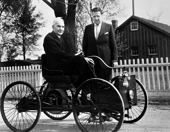 A black and white photo showing an elderly Henry Ford seated in a very early Ford car, with Henry Ford II standing alongside.