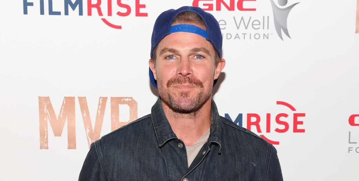 stephen amell stands looking at the camera with a neutral facial expression, he has brown hair and beard, he wears a cap backwards, grey tshirt, blue denim jacket and jeans