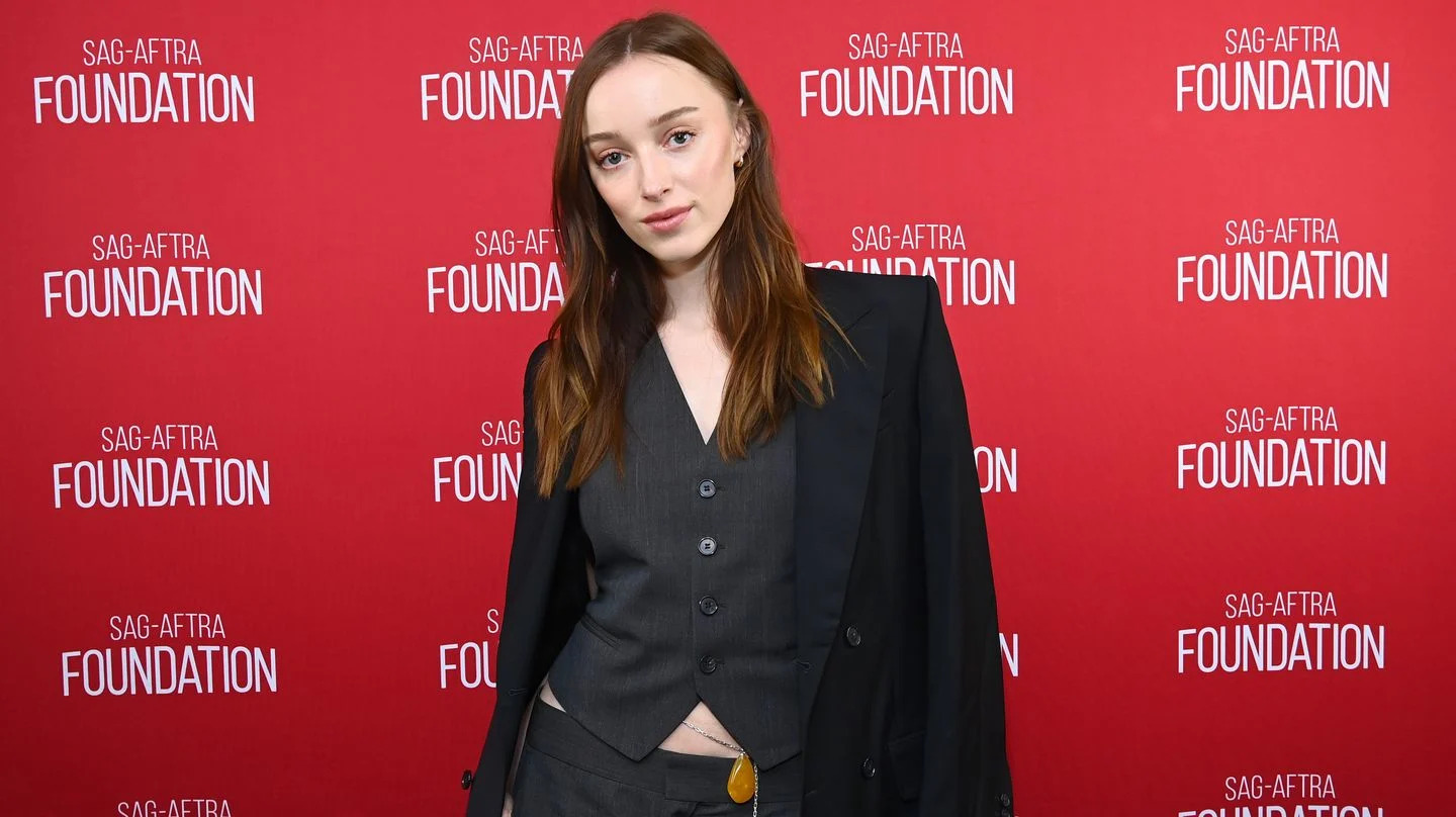 phoebe dynevor, a young woman stands looking at the camera with a neutral facial expression, she has long brown hair worn in loose waves, she wears a dark grey waistcoat and skirt with a black blazer