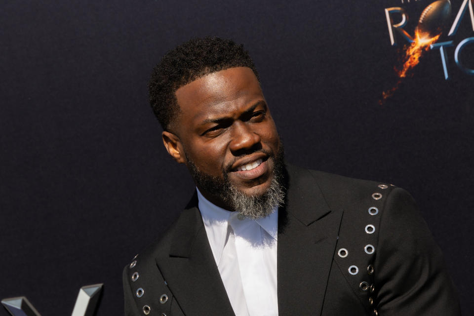 Kevin Hart wearing a stylish black suit with grommet detailing at the premiere event of "The Road to" against a dark backdrop